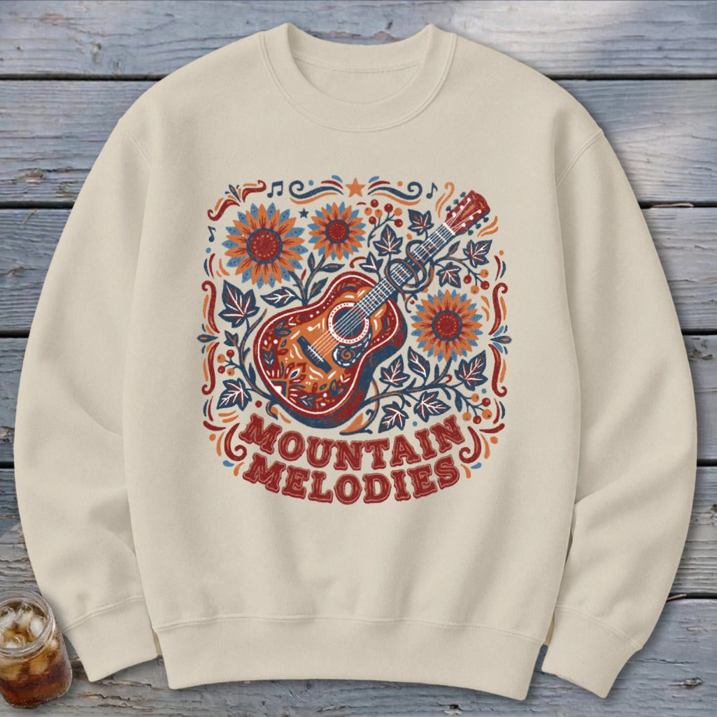 A cream-colored crewneck sweatshirt featuring a graphic of an acoustic guitar, sunflowers, and the text "MOUNTAIN MELODIES".