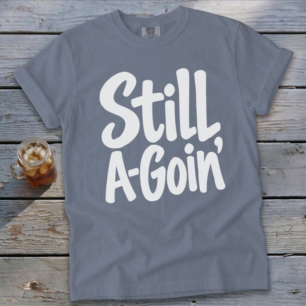 A slate gray short-sleeve t-shirt with white handwritten text reading Still A-Goin' on a wooden surface.