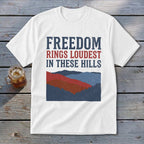 A white t-shirt featuring the distressed text FREEDOM RINGS LOUDEST IN THESE HILLS above a red, white, and blue mountain graphic.