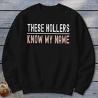 Black sweatshirt with white text "THESE HOLLERS" and beige text "KNOW MY NAME".