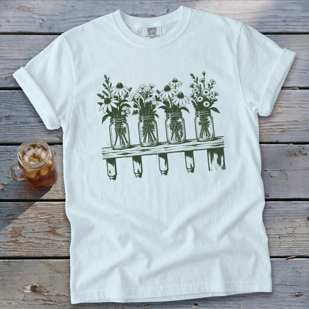 A white t-shirt featuring a green graphic of four mason jars filled with wildflowers on a shelf.