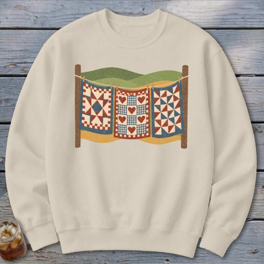 Cream crewneck sweatshirt featuring a graphic of three vintage quilts hanging on a clothesline outdoors.