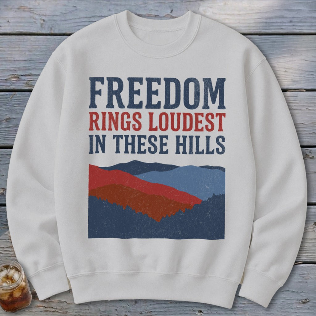 Off-white crewneck sweatshirt featuring blue and red text that reads FREEDOM RINGS LOUDEST IN THESE HILLS above a mountain landscape graphic.