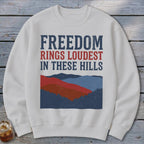 Off-white crewneck sweatshirt featuring blue and red text that reads FREEDOM RINGS LOUDEST IN THESE HILLS above a mountain landscape graphic.