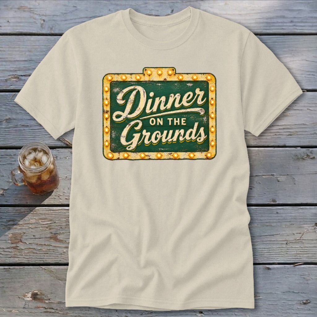 A cream-colored crewneck t-shirt featuring a green vintage sign graphic with the text Dinner on the Grounds.