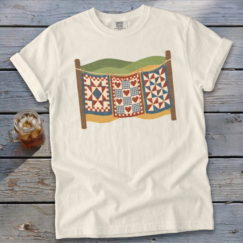 A cream-colored t-shirt with a graphic of three colorful quilts hanging on a clothesline.