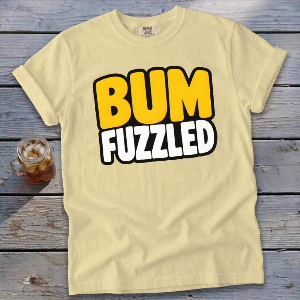 A light yellow t-shirt with rolled sleeves features the text "BUM FUZZLED" centered on the chest, displayed on a weathered wooden surface next to a glass of iced tea.