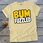 A light yellow t-shirt with rolled sleeves features the text "BUM FUZZLED" centered on the chest, displayed on a weathered wooden surface next to a glass of iced tea.