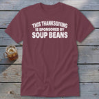 Maroon crewneck t-shirt with white text reading "This Thanksgiving is sponsored by soup beans" laid flat on a rustic wooden surface.