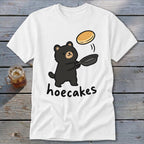 A white t-shirt featuring a cartoon black bear flipping a pancake with the text hoecakes.