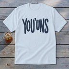 White t-shirt with "YOU'UNS" in dark blue lettering, on a wooden background.