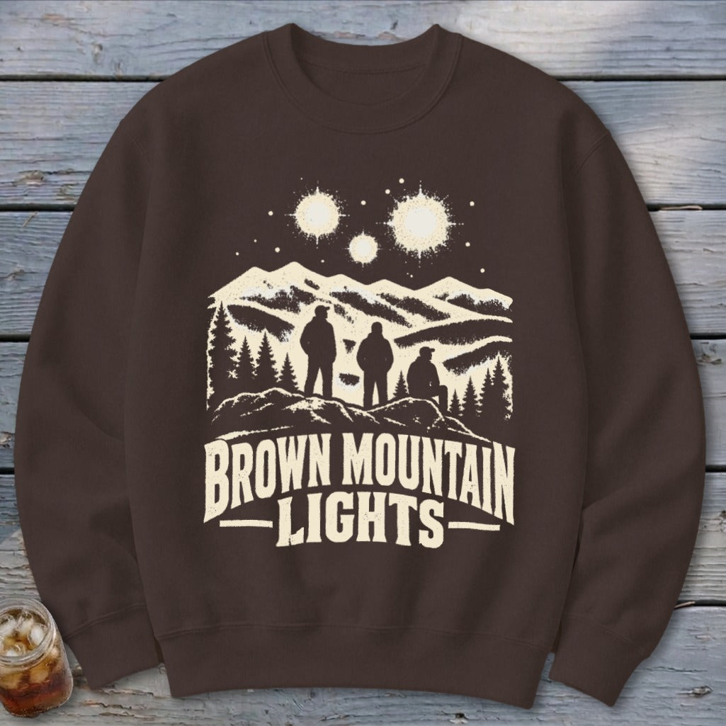 Brown crewneck sweatshirt featuring a graphic of three figures, mountains, glowing lights, and the text Brown Mountain Lights.