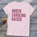 Pink t-shirt with "NORTH CAROLINA RAISED" in maroon text.