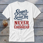 A white t-shirt featuring blue and red text that reads Sugar in my Sweat Tea NEVER IN MY CORNBREAD, laid on a wooden surface with a glass of iced tea nearby.