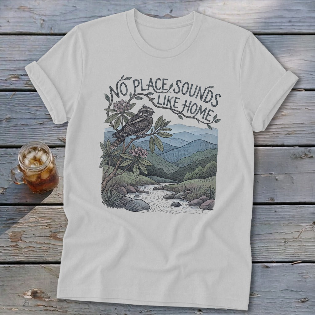 White t-shirt with "No Place Sounds Like Home" text, bird on branch, and mountain stream landscape.