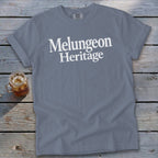 A slate blue t-shirt printed with the white text Melungeon Heritage lies on a weathered wooden surface beside a glass of iced tea.