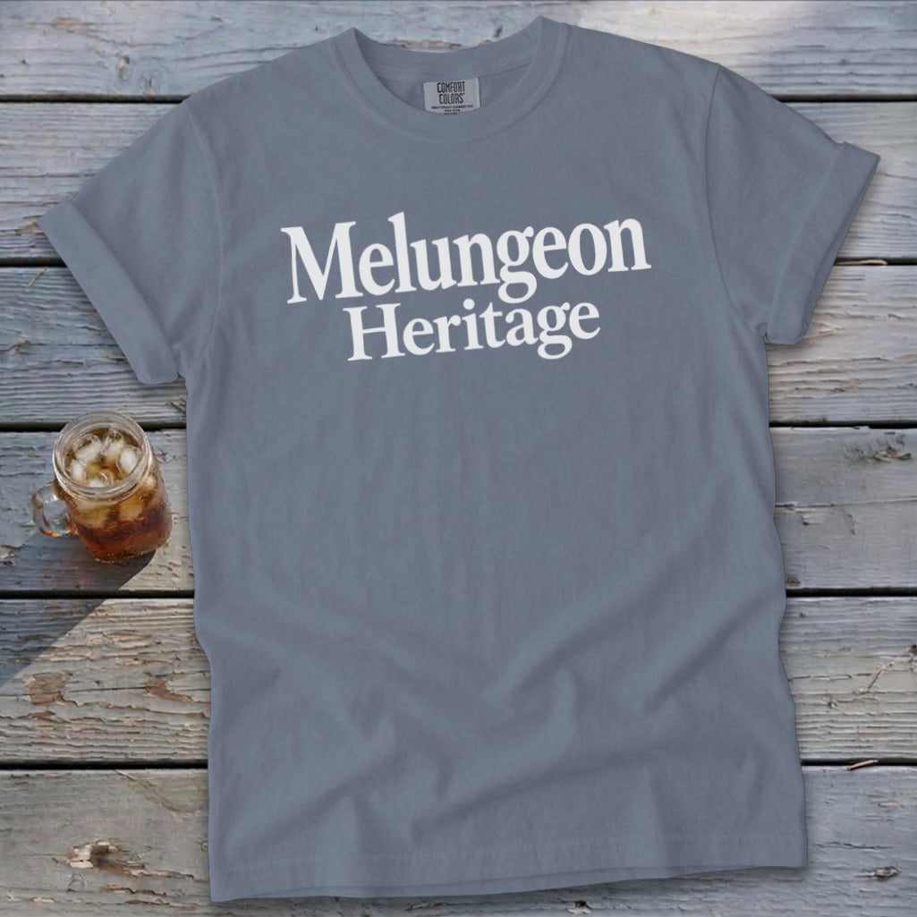 A slate blue t-shirt printed with the white text Melungeon Heritage lies on a weathered wooden surface beside a glass of iced tea.