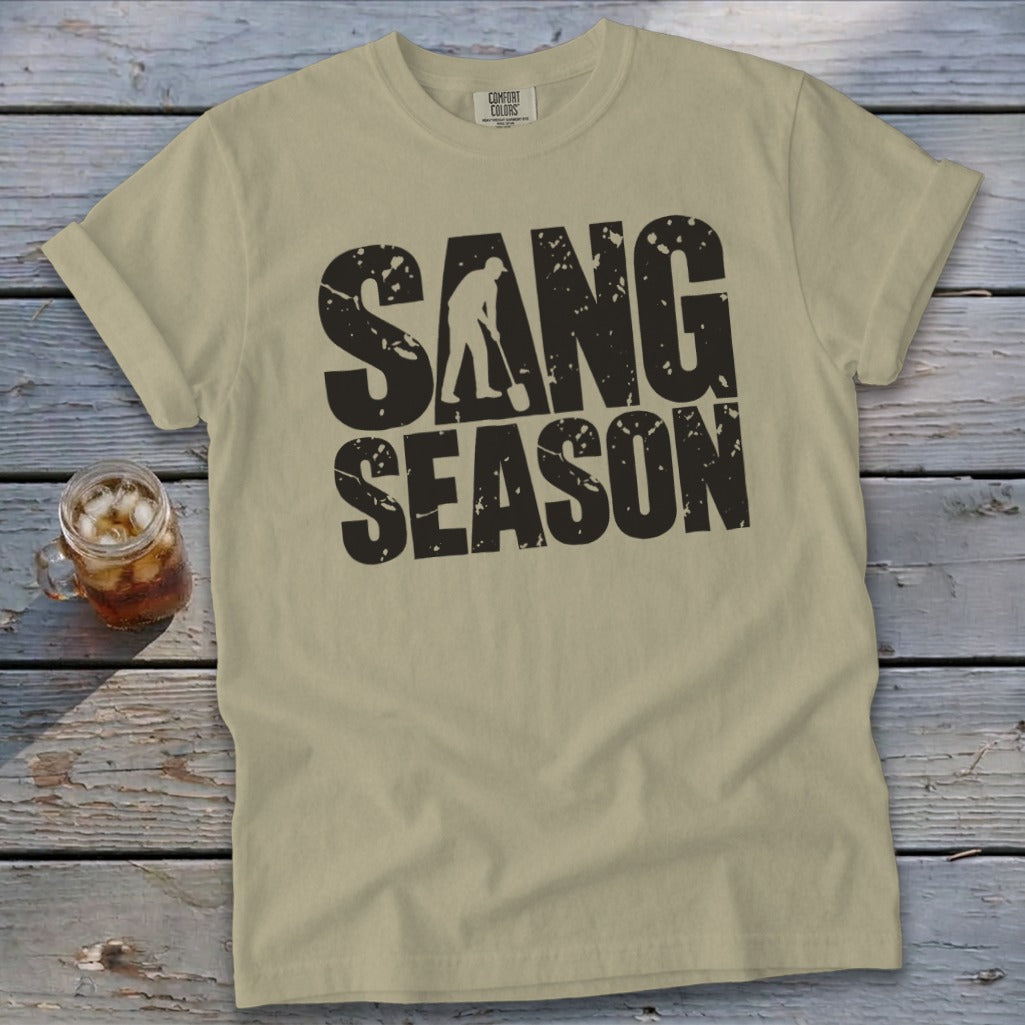 Khaki t-shirt featuring bold black distressed text reading SANG SEASON with a silhouette of a ginseng digger inside the letter N.