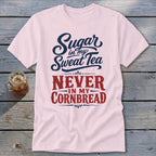 Light pink t-shirt featuring blue and red text that reads Sugar in my Sweat Tea Never in my Cornbread laid on a wooden table.