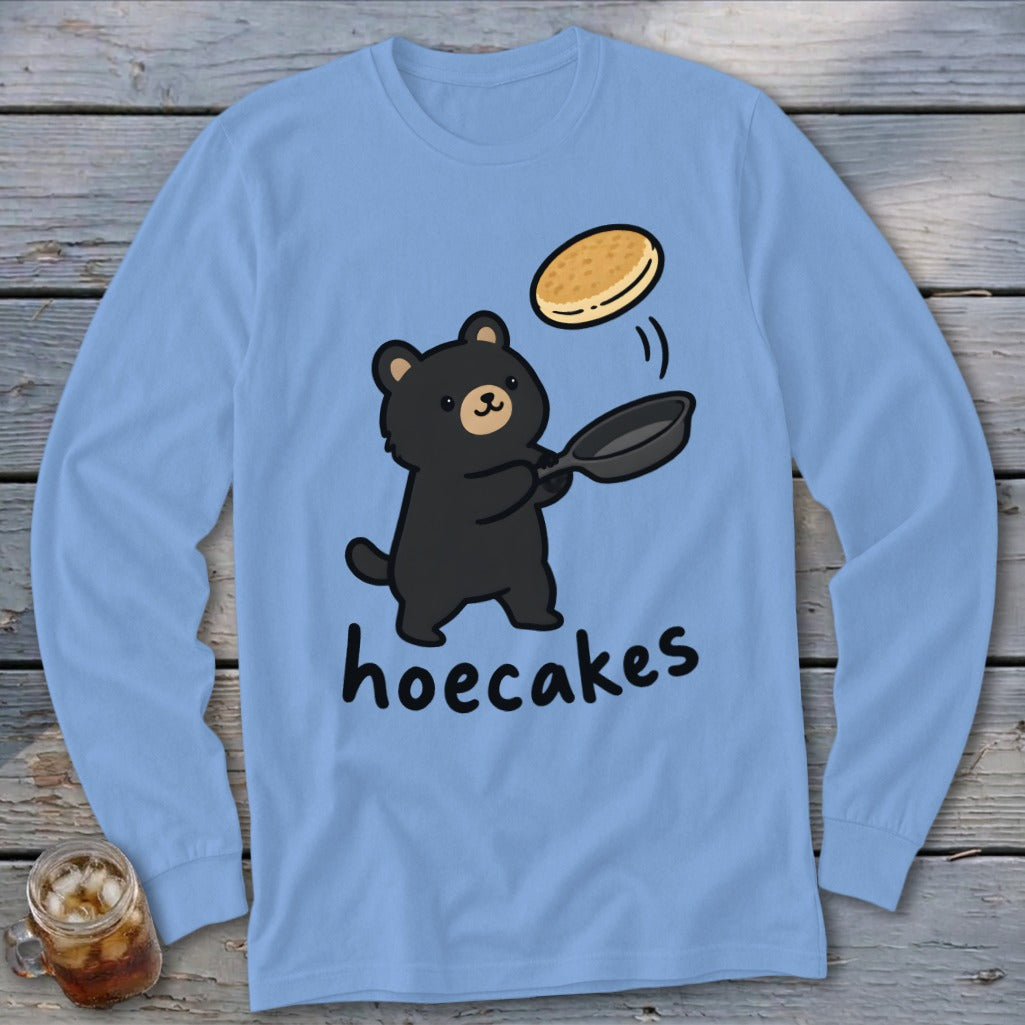 A light blue long-sleeve t-shirt featuring a cartoon black bear flipping a pancake from a frying pan above the word hoecakes.