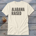 Cream t-shirt with black "ALABAMA RAISED" text on wooden background.