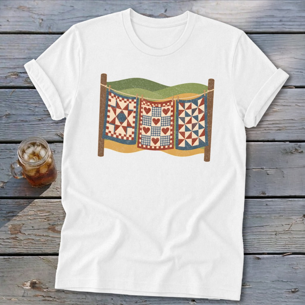 A white t-shirt featuring a graphic of three patchwork quilts hanging on a clothesline against a landscape.