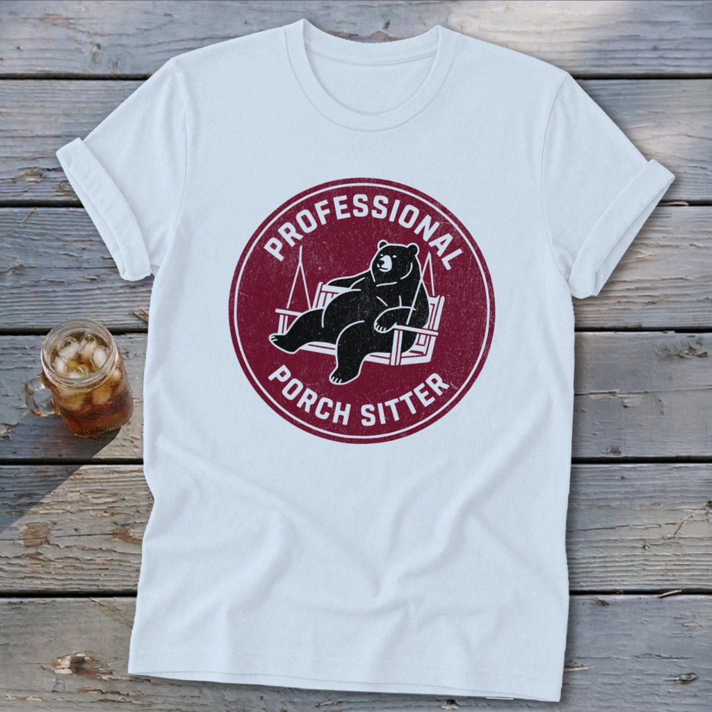 A white t-shirt featuring a maroon graphic of a bear lounging on a porch swing, surrounded by the text PROFESSIONAL PORCH SITTER.