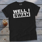 A black t-shirt with the white text "WELL I SWAN" on a wooden surface next to a glass of iced tea.