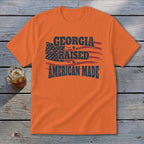 An orange crewneck t-shirt featuring a distressed American flag and text that reads Georgia Raised American Made.