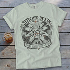 T-shirt design with hands sewing a quilt and the text "STITCHED BY KIN EVERY SQUARE TELLS A STORY".