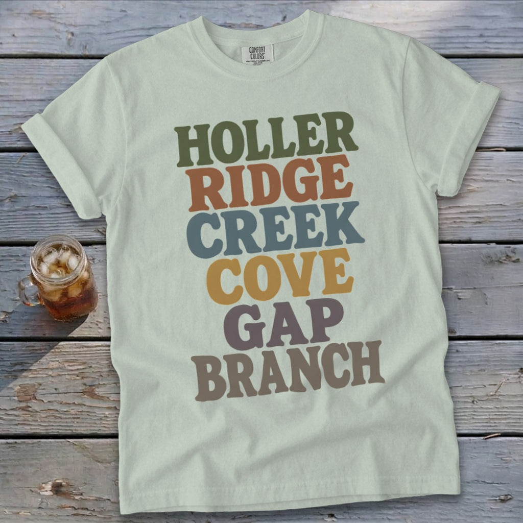 A light green t-shirt with stacked multicolored text reading HOLLER RIDGE CREEK COVE GAP BRANCH rests on wooden boards.