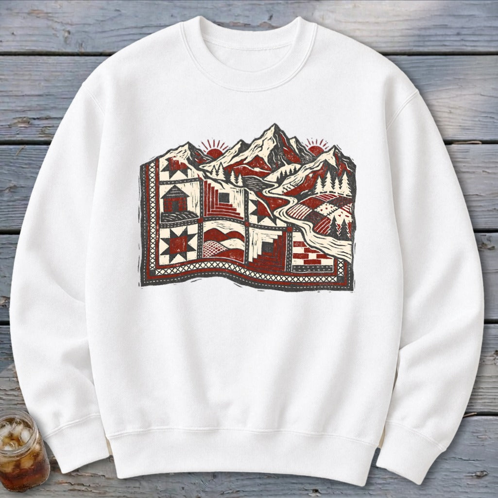 A white crewneck sweatshirt featuring a graphic print of a quilt layered over a mountain landscape.