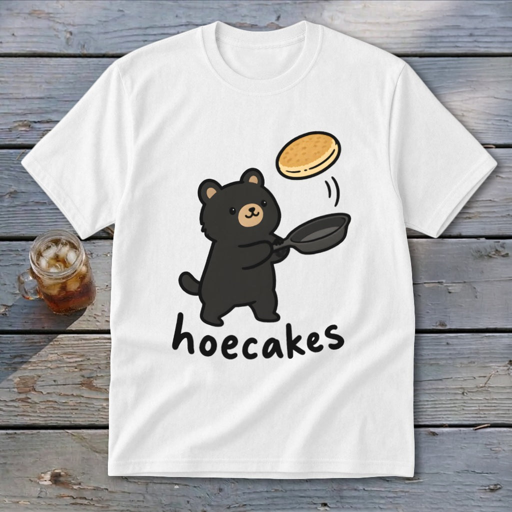 A white t-shirt featuring a cartoon black bear flipping a pancake from a skillet, with the text hoecakes underneath.