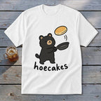 A white t-shirt featuring a cartoon black bear flipping a pancake from a skillet, with the text hoecakes underneath.