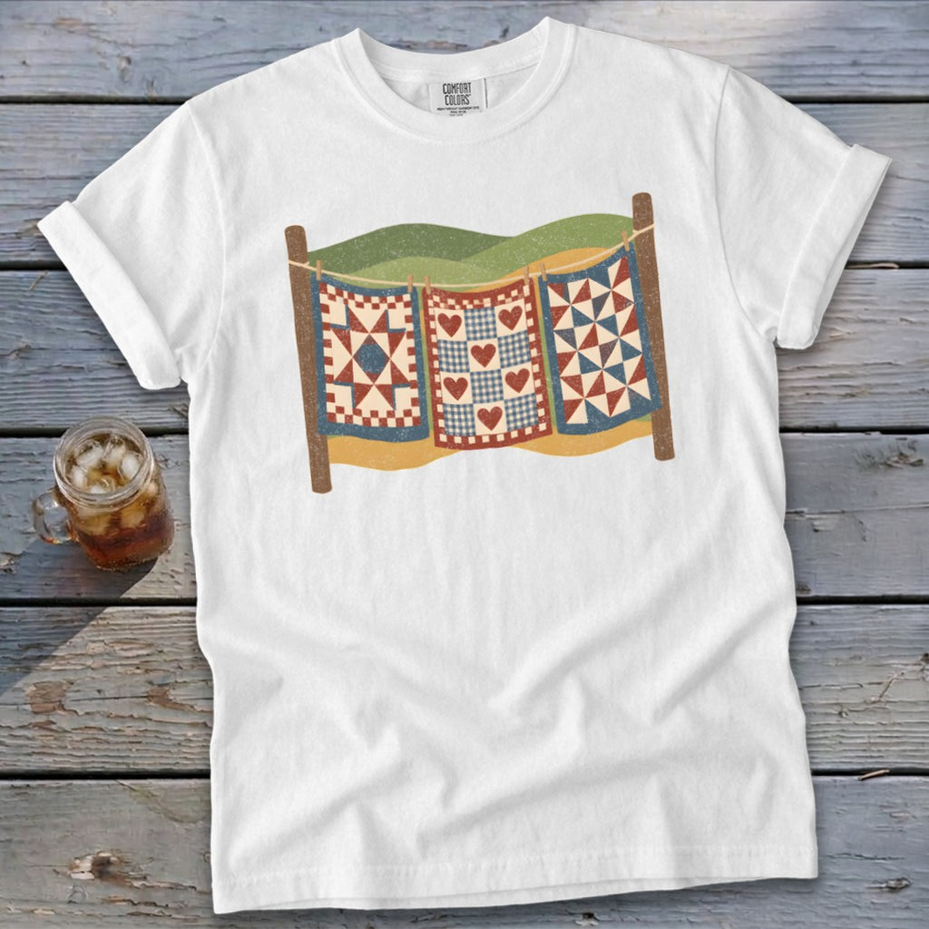 A white crewneck t-shirt featuring a graphic of three patchwork quilts hanging on a clothesline.