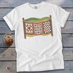 A white crewneck t-shirt featuring a graphic of three patchwork quilts hanging on a clothesline.