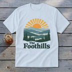 Light blue t-shirt with sunset over mountains and "The Foothills" text.