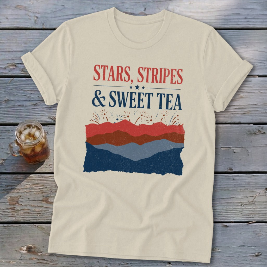 A cream t-shirt rests on a wooden surface, featuring the red and blue text Stars Stripes & Sweet Tea above red, white, and blue layered mountain and firework graphics.
