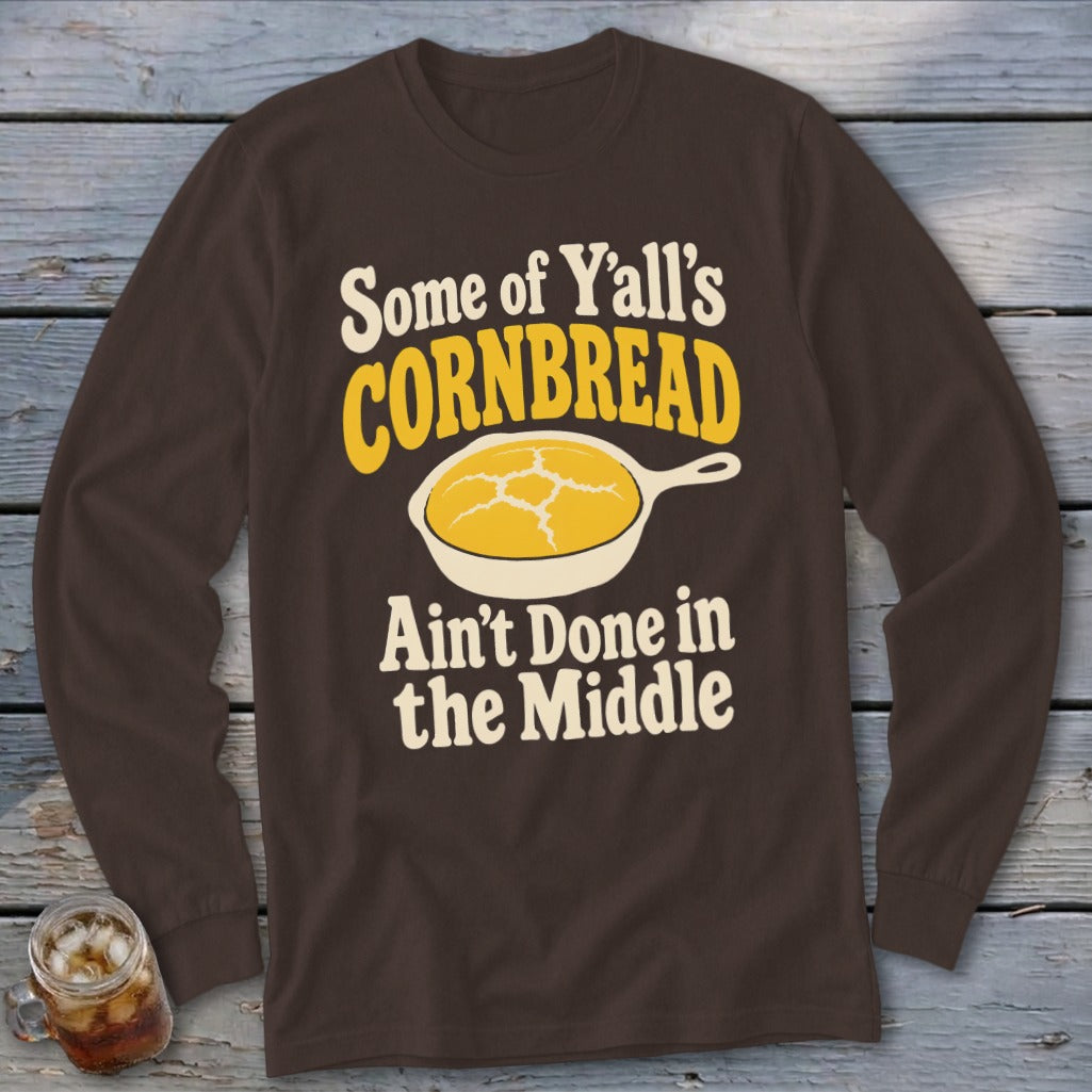 A brown long-sleeved shirt with the yellow and white text "Some of Y'all's Cornbread Ain't Done in the Middle" and an illustration of cornbread in a skillet.
