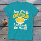 Turquoise t-shirt with rolled sleeves displaying text and a cornbread graphic on a rustic wooden background.