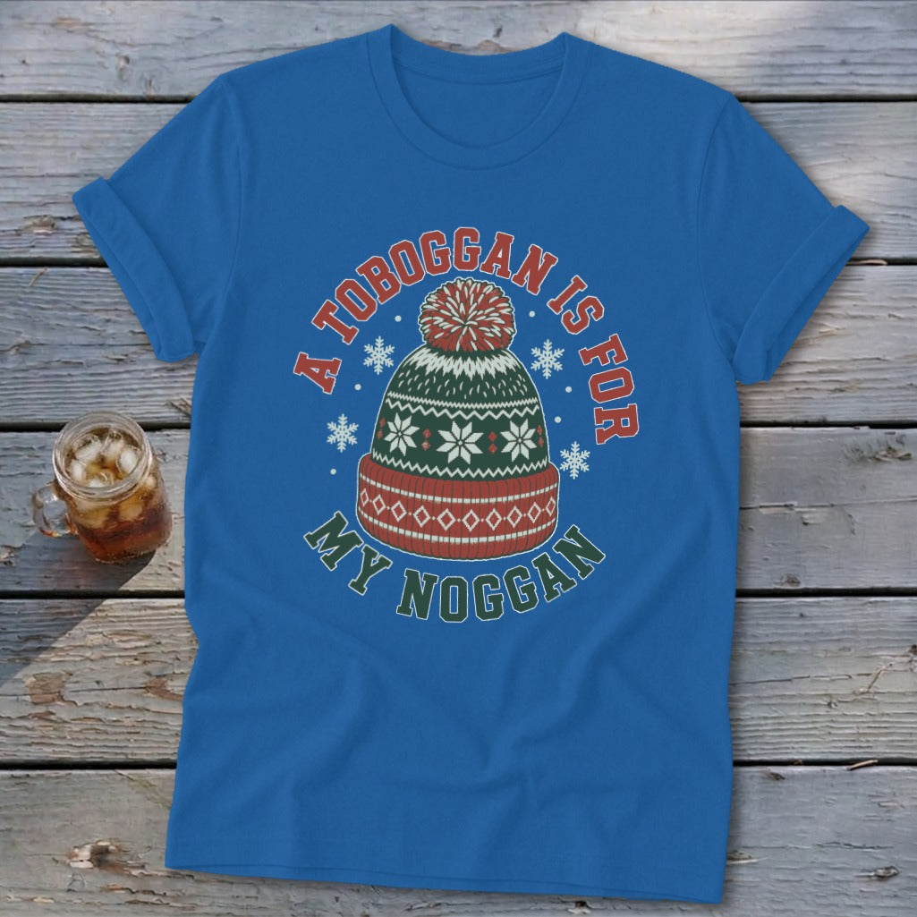 A blue t-shirt featuring a winter graphic and text reading A TOBOGGAN IS FOR MY NOGGAN on a wooden surface.