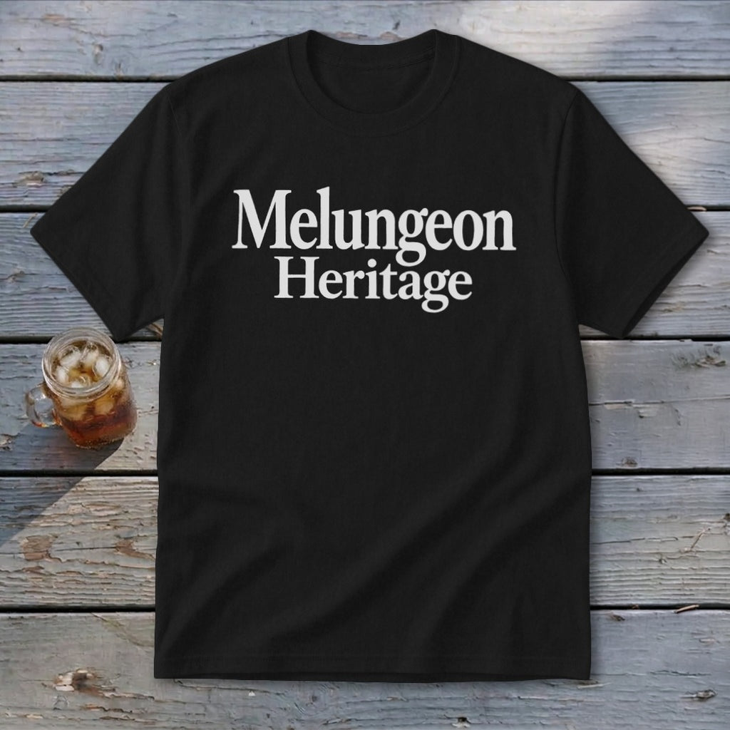 A black t-shirt printed with the white text Melungeon Heritage rests on weathered wooden planks beside a mason jar of iced tea.