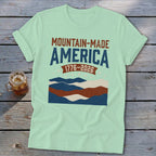Light green t-shirt featuring distressed Mountain-Made America and 1776-2026 text with layered mountain graphics.