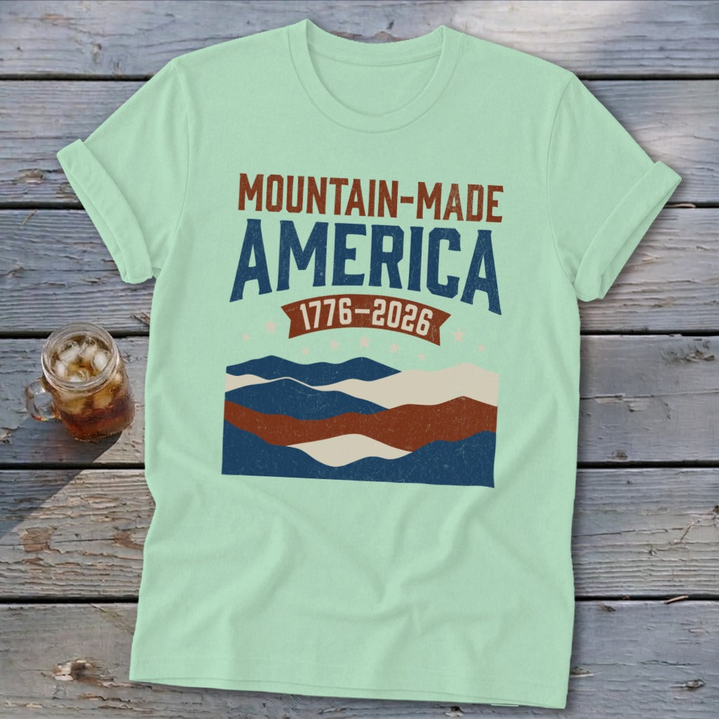 Light green t-shirt featuring distressed Mountain-Made America and 1776-2026 text with layered mountain graphics.