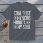 A dark gray t-shirt featuring the distressed white text, COAL DUST IN MY VEINS MOUNTAINS IN MY SOUL, rests on a weathered wooden surface next to a glass of iced tea.