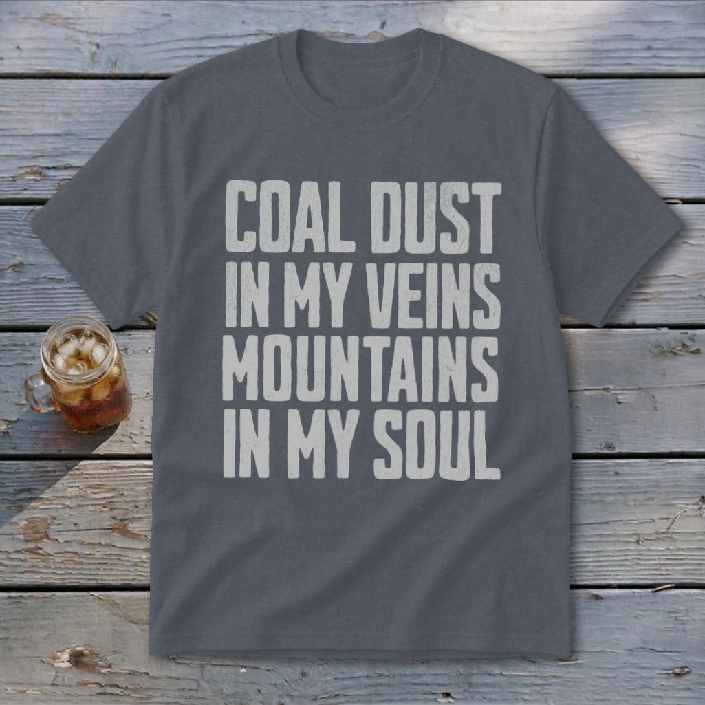 A dark gray t-shirt featuring the distressed white text, COAL DUST IN MY VEINS MOUNTAINS IN MY SOUL, rests on a weathered wooden surface next to a glass of iced tea.