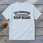 A light blue t-shirt with black text reading "THIS THANKSGIVING IS SPONSORED BY SOUP BEANS" lies on a wooden background.