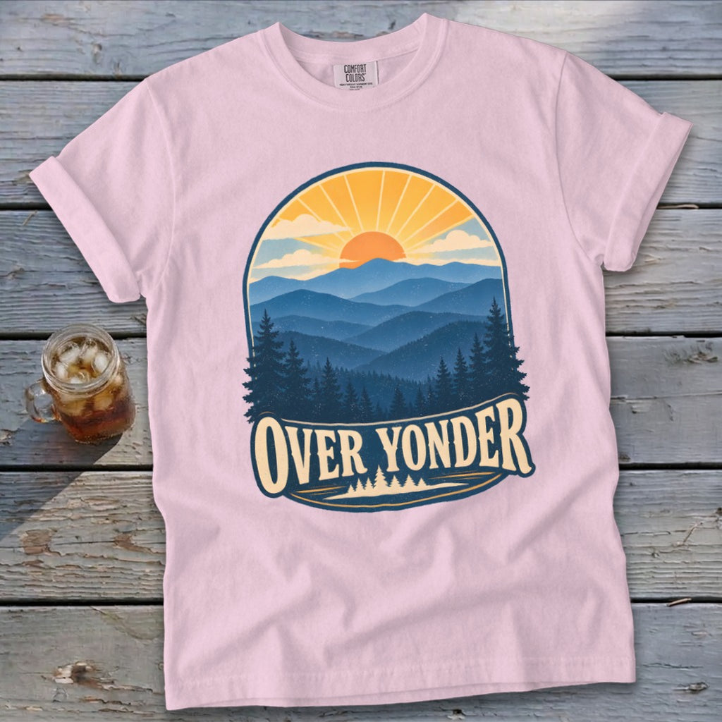 Light pink t-shirt featuring a mountain and sunrise graphic with the text Over Yonder, laid on wooden planks.