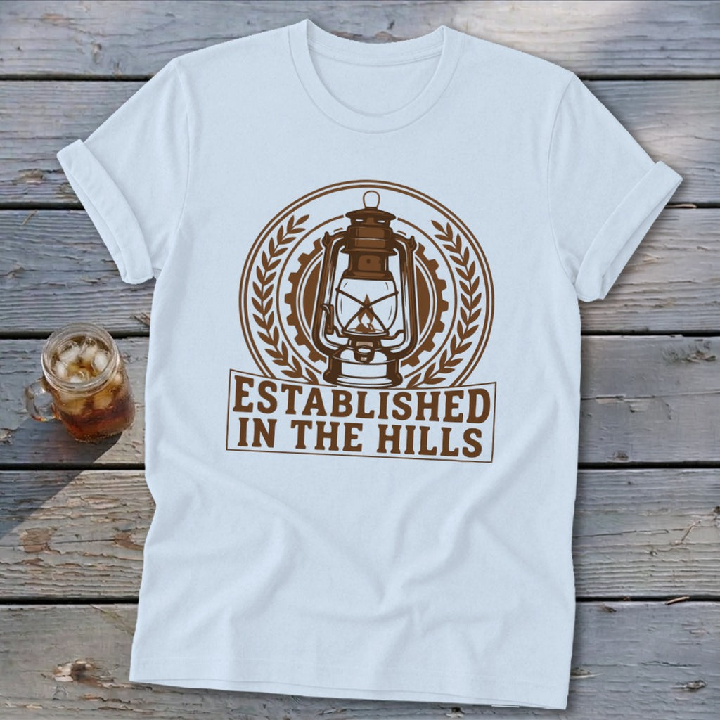White t-shirt with brown circular graphic of a lantern and text "Established in the Hills".