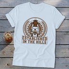 White t-shirt with brown circular graphic of a lantern and text "Established in the Hills".